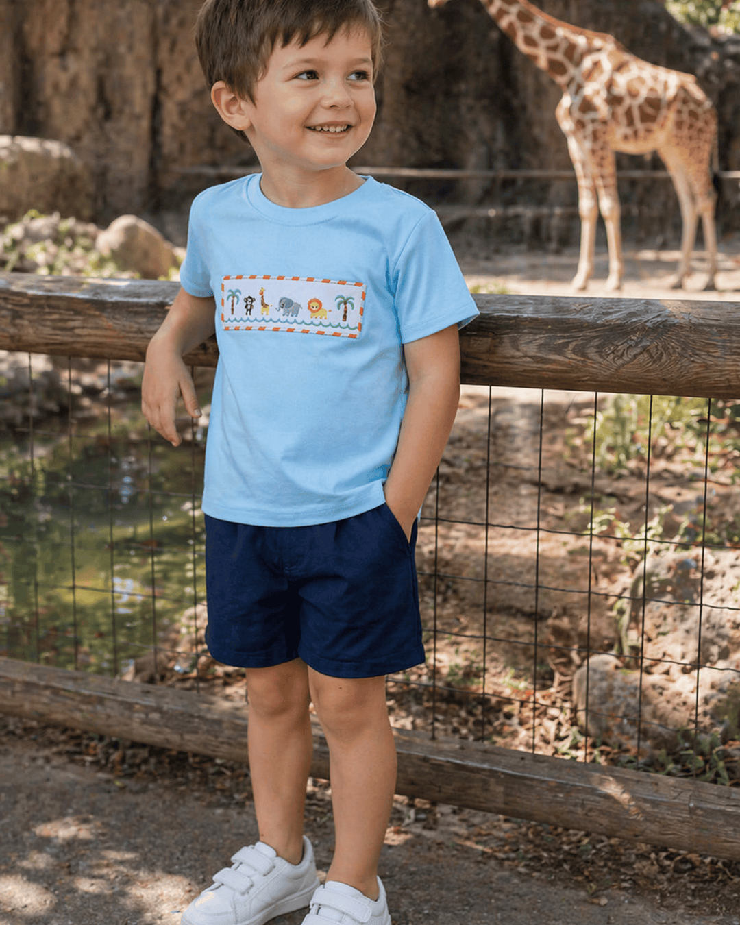 Child wearing a light blue t-shirt with a colorful design and navy shorts, standing in front of a wooden fence with a giraffe in the background.