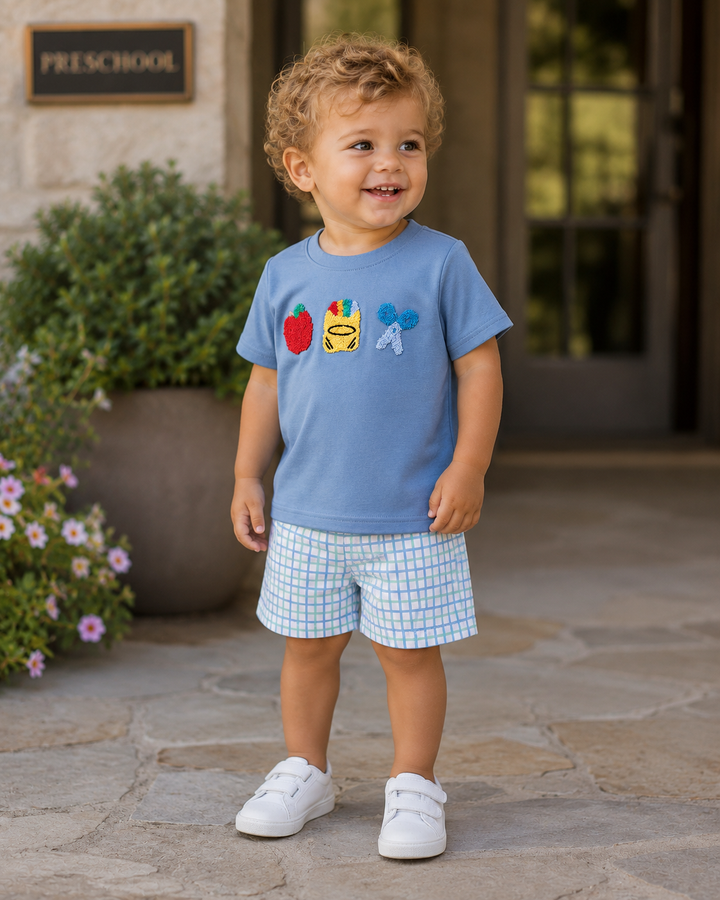 A toddler boy wearing a light blue short-sleeve t-shirt with colorful fruit graphics and light blue and white checkered shorts, standing on a stone patio in front of a building entrance.