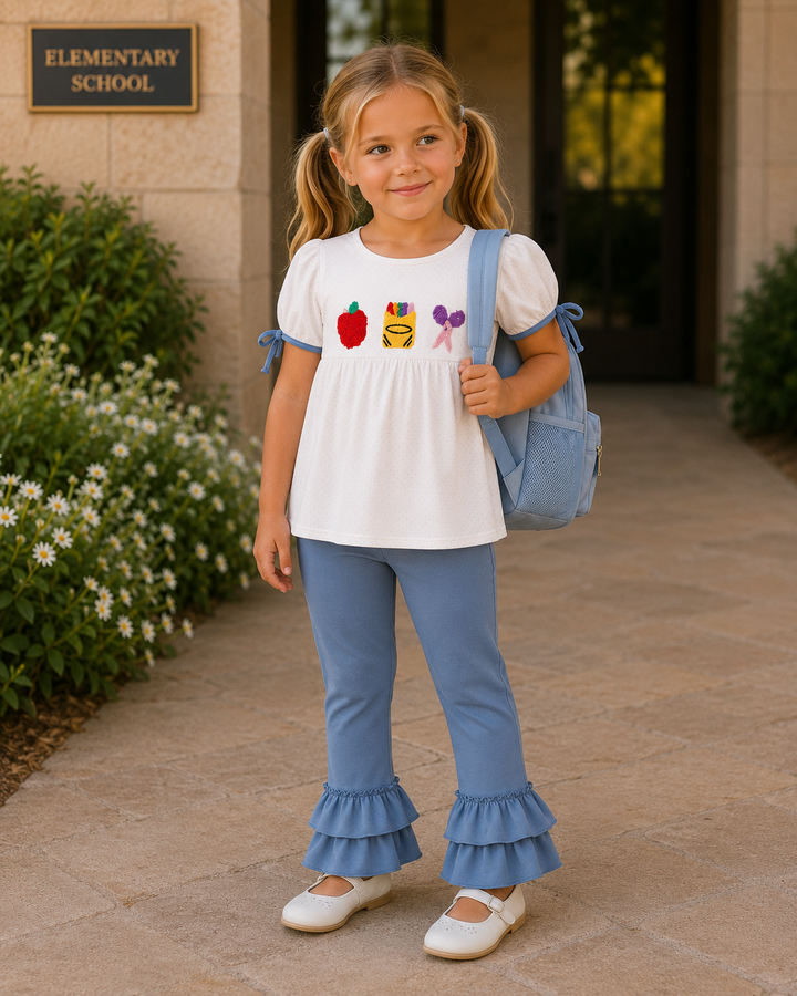 A young girl stands on a school walkway wearing a white short-sleeve top with colorful school-themed graphics on the chest, light blue ruffled flare pants, and white shoes, while carrying a light blue backpack.