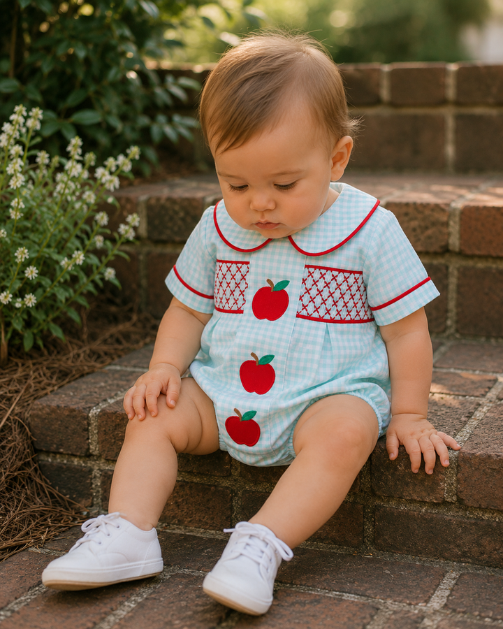 Baby wearing a light blue romper with red apple patterns, sitting on brick steps.