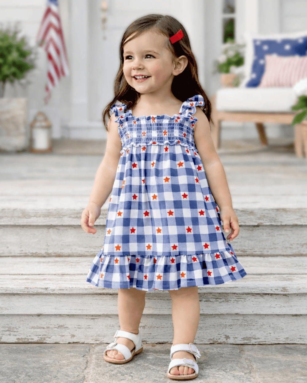 Young girl in a blue checkered dress with red and white stars standing on a wooden porch.