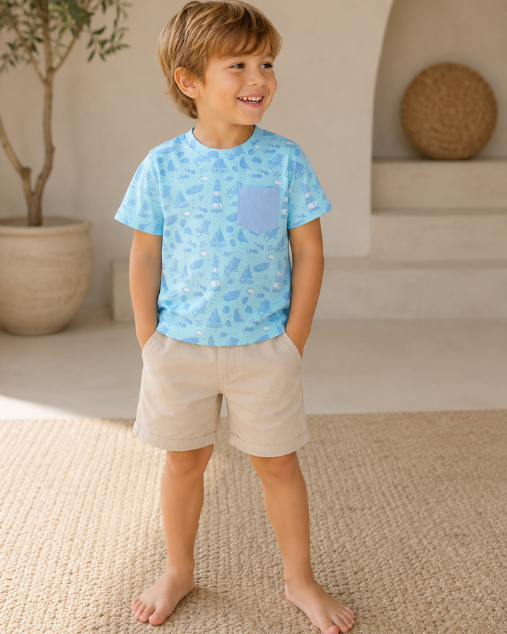 Young boy wearing a blue patterned shirt and beige shorts standing in a room with a plant and woven basket in the background.
