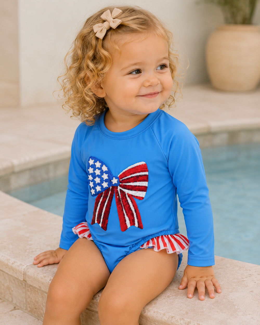 Child wearing a blue swimsuit with an American flag bow design by a pool.