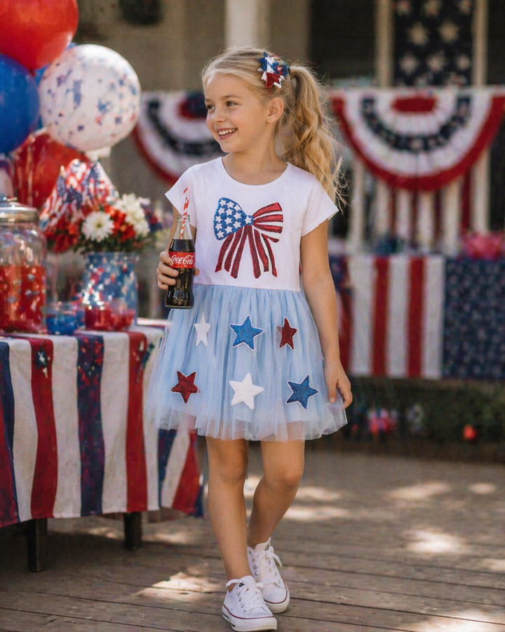 Young girl in a patriotic outfit with a dress featuring stars and a bow, holding a Coca-Cola bottle, standing in front of a decorated outdoor setting.