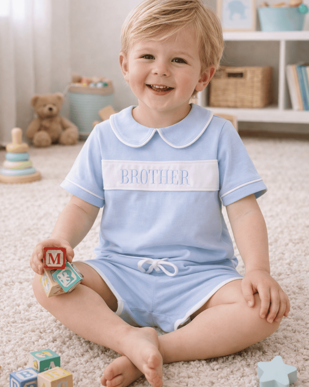 Child wearing a light blue 'BROTHER' onesie sitting on a carpeted floor with toys around.