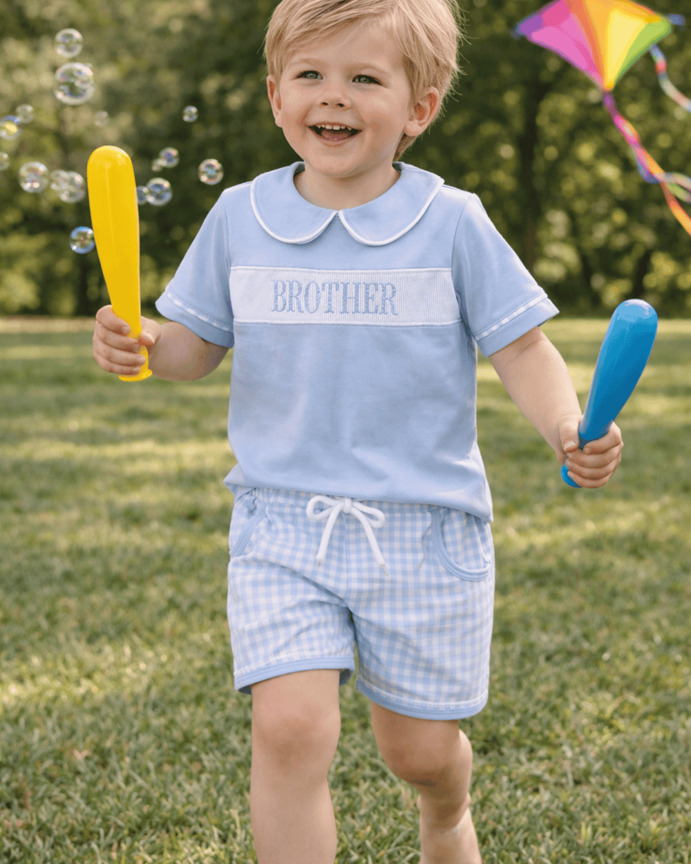 Child wearing a 'BROTHER' shirt holding two bat toys outdoors with bubbles and a kite in the background.