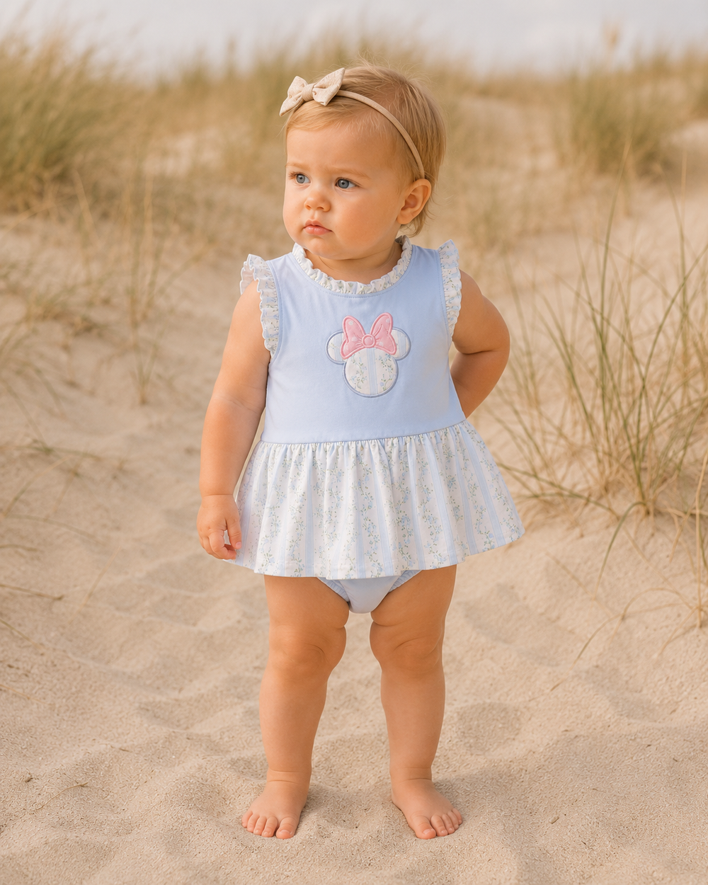 Baby girl in a light blue swimsuit with ruffles standing on sandy dunes.