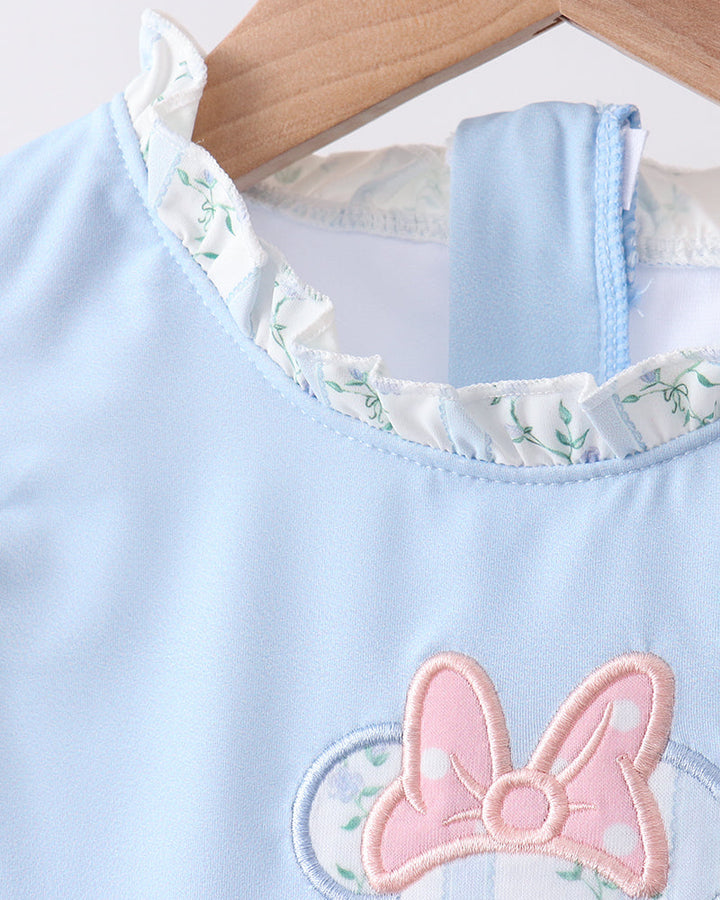 Light blue swim top with a pink bow and floral trim on a wooden hanger.