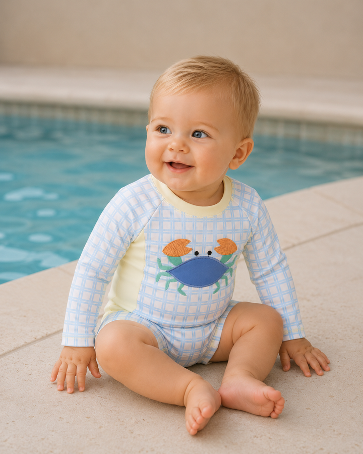 Baby wearing a blue checkered swimsuit with a crab design by a pool.