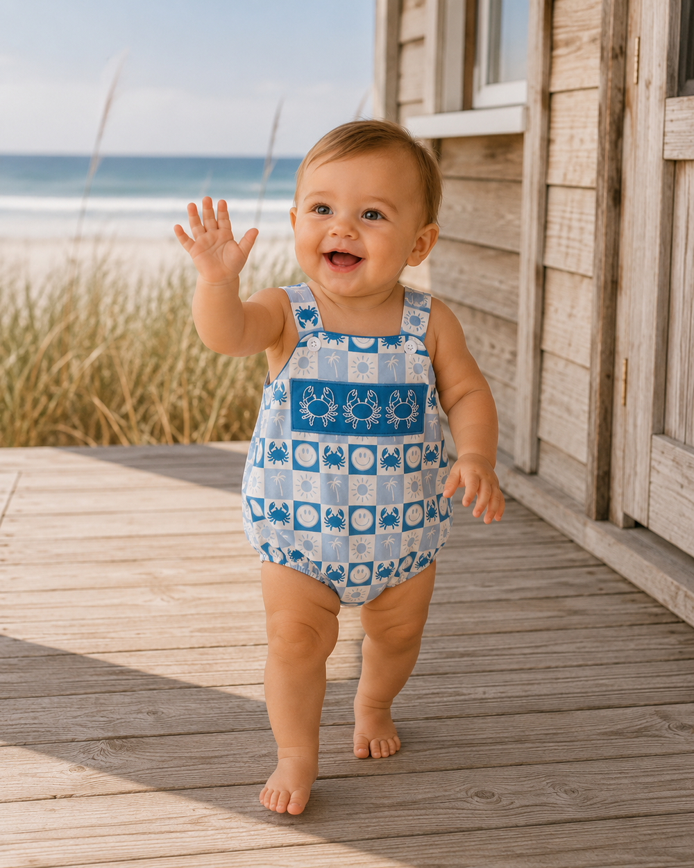A baby walking barefoot on a wooden deck next to the ocean, wearing a blue and white patterned sleeveless romper with sea-themed squares.