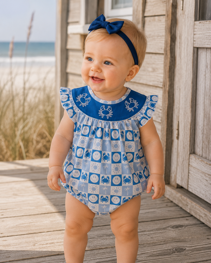 A baby girl wearing a blue and white patterned romper with a matching blue bow headband, standing on a wooden deck near the beach.