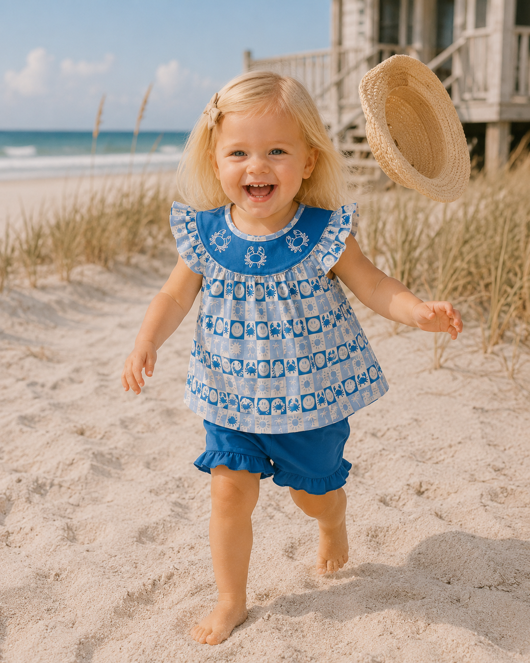 A young blonde girl smiling and running on a sandy beach, wearing a blue and white patterned ruffle-sleeve top and matching blue ruffle shorts, with a straw hat floating near her head.