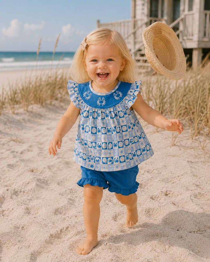 A young blonde girl smiling and running on a sandy beach, wearing a blue and white patterned ruffle-sleeve top and matching blue ruffle shorts, with a straw hat floating near her head.