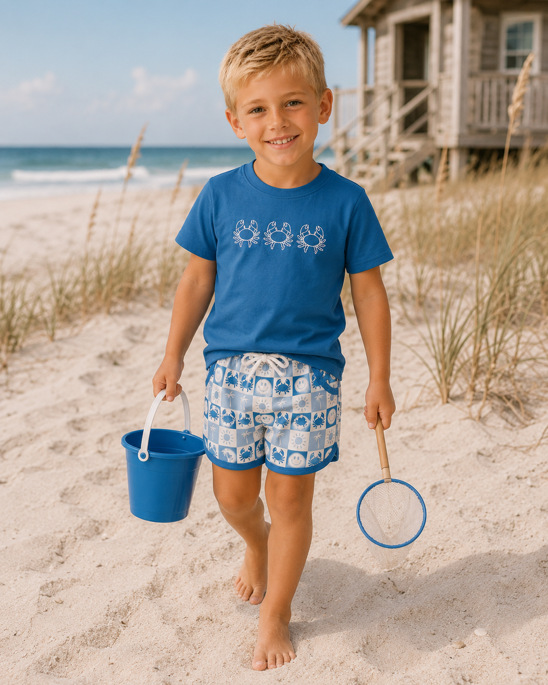 A young boy walking on a sandy beach wearing a blue t-shirt with a white octopus pattern and matching blue-and-white patterned swim trunks, holding a blue bucket and a net. A beach house and ocean are visible in the background.