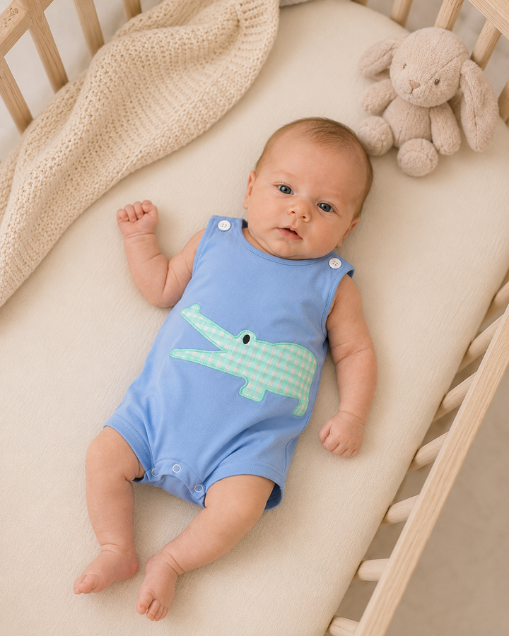 A baby lying on a cream mattress in a wooden crib, wearing a light blue sleeveless romper with a green and white gingham crocodile graphic on the front. A beige knit blanket and a plush bunny toy are visible in the crib.