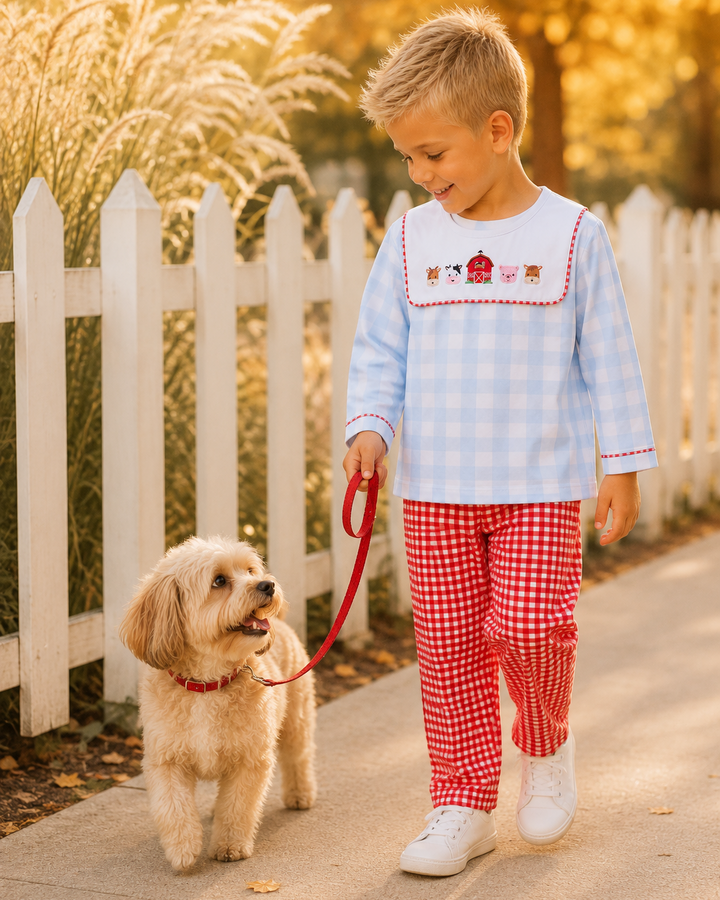 A young boy walking a small fluffy dog on a red leash outdoors. The boy is wearing a light blue and white gingham long-sleeve shirt with red trim and embroidered details on the chest, paired with red and white gingham pants.