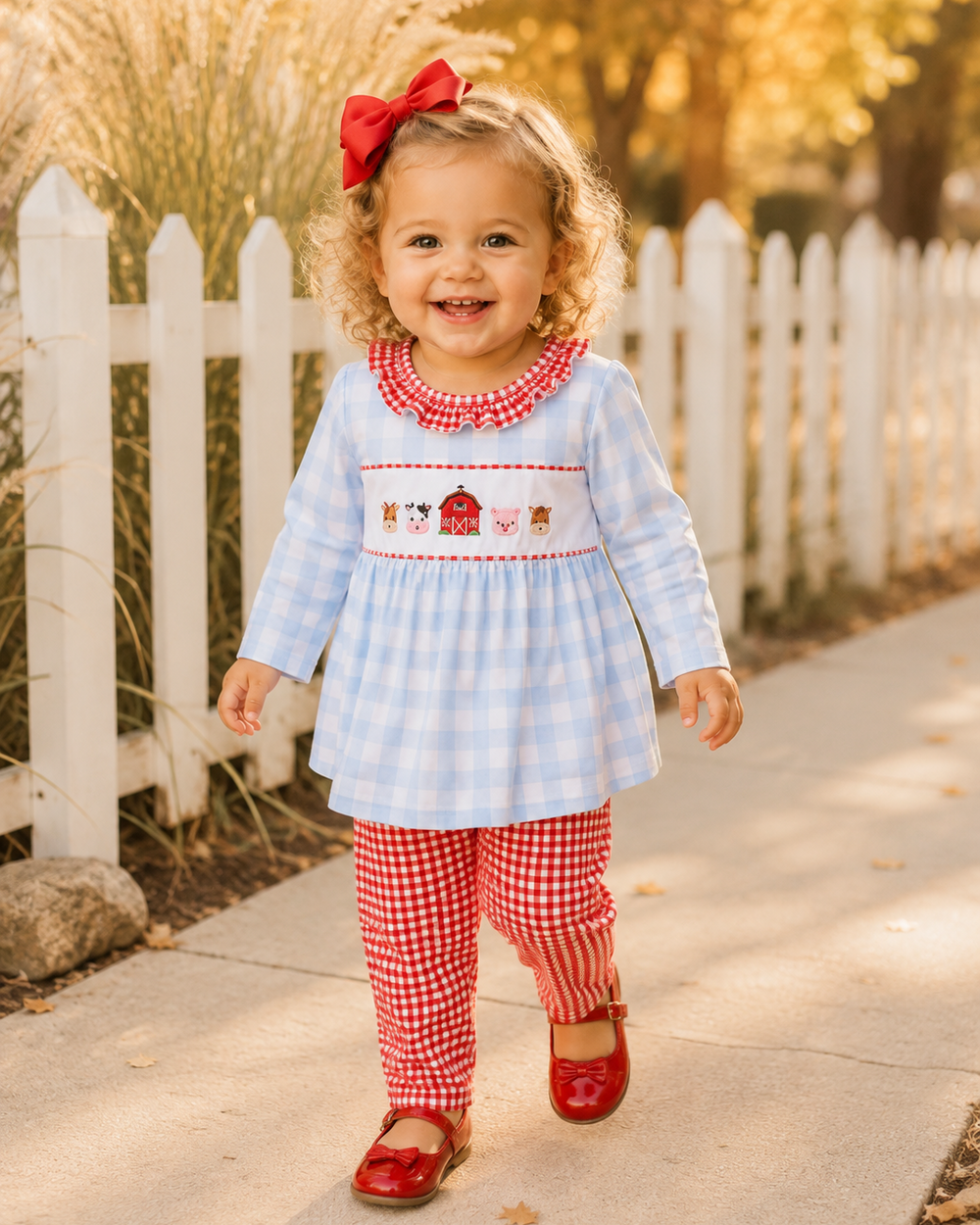 A toddler girl walking outdoors on a sunlit sidewalk beside a white picket fence. She is wearing a light blue and white gingham dress with red trim and a smocked chest panel featuring a barn motif, paired with red-and-white gingham leggings and red flats.