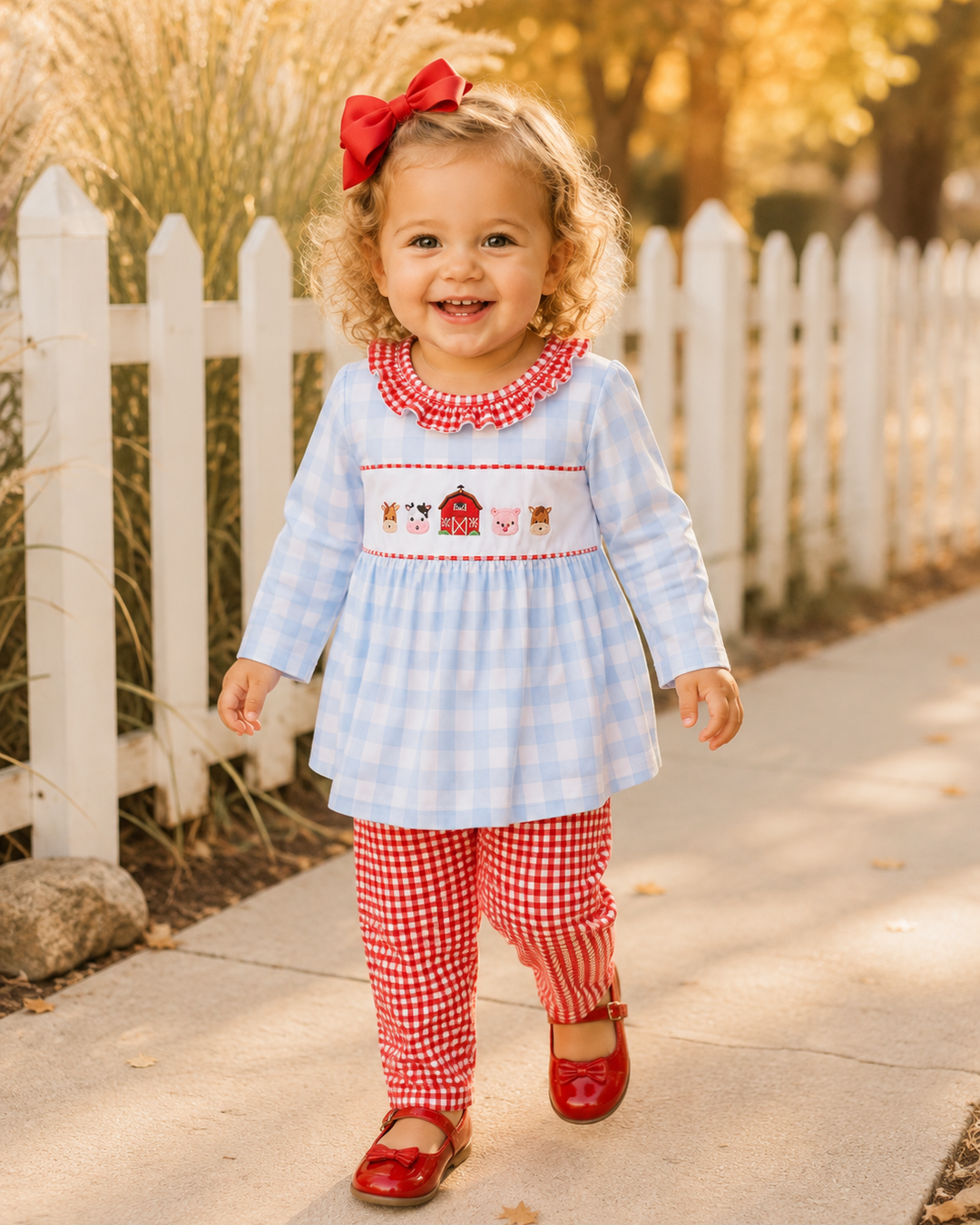A toddler girl walking outdoors on a sunlit sidewalk beside a white picket fence. She is wearing a light blue and white gingham dress with red trim and a smocked chest panel featuring a barn motif, paired with red-and-white gingham leggings and red flats.