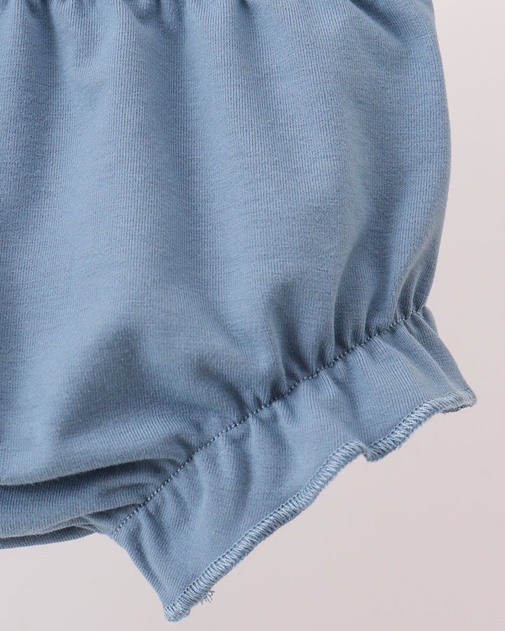 Close-up of a light blue fabric garment with gathered elastic waistband and ruffled hem, shown against a pale pink background.