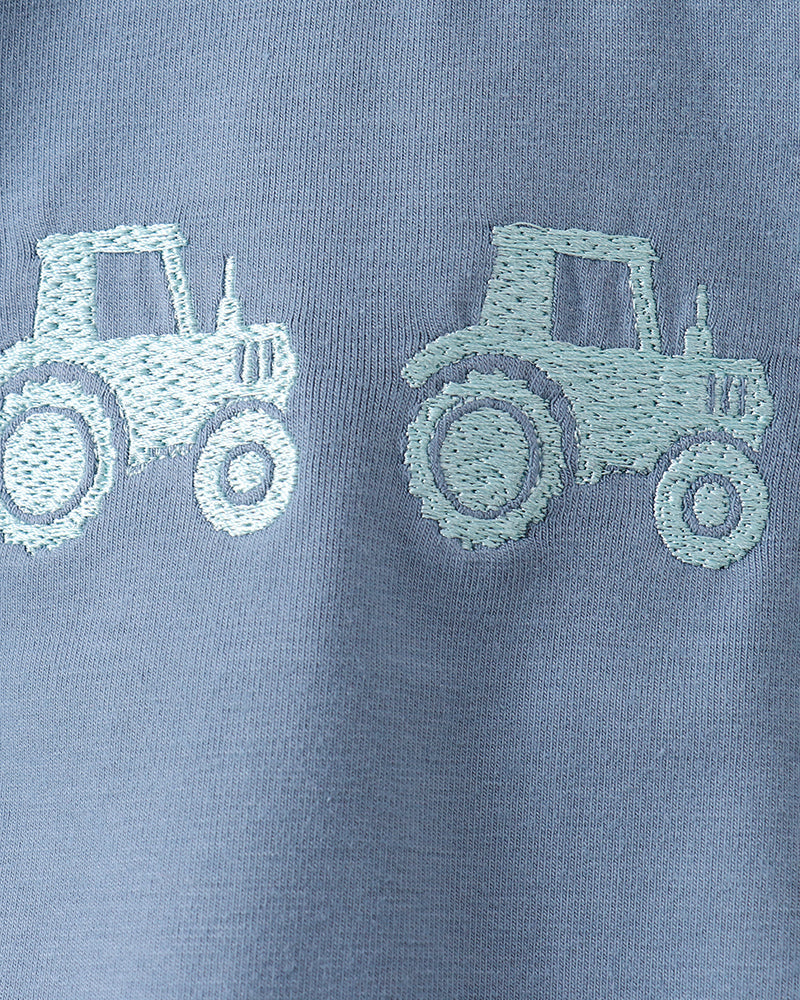 Close-up of a light blue fabric garment with two pale mint-green embroidered tractor motifs on the chest.