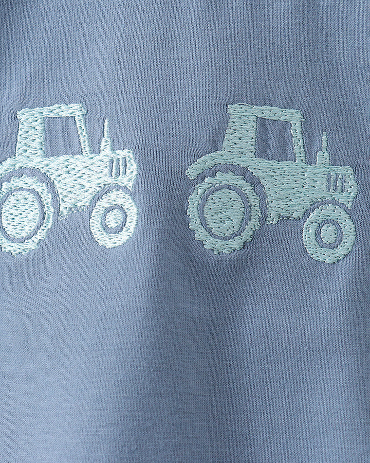 Close-up of a light blue fabric garment with two pale mint-green embroidered tractor motifs on the chest.