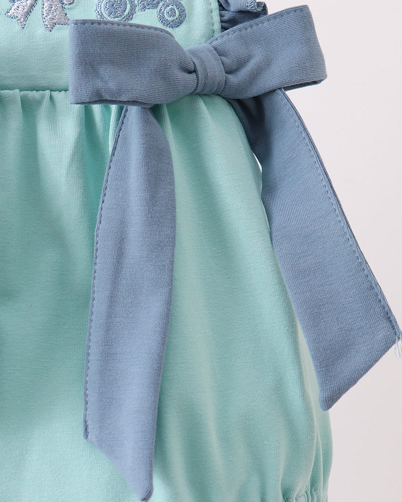 Close-up of a mint green baby romper with a dusty blue bow on the front, featuring embroidered tractors and bows on the chest.