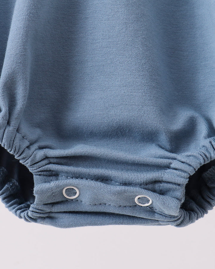 Close-up of a dusty blue baby romper or bodysuit showing the elasticized crotch area with two silver metal snap buttons on a light gray background.