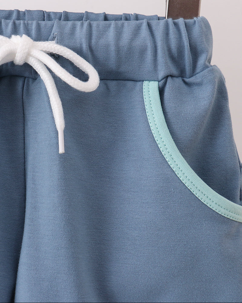 Close-up of dusty blue shorts hanging on a wooden hanger, featuring a white drawstring and a mint green contrast trim along the pocket edge.