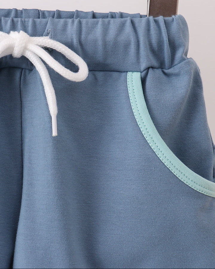 Close-up of dusty blue shorts hanging on a wooden hanger, featuring a white drawstring and a mint green contrast trim along the pocket edge.