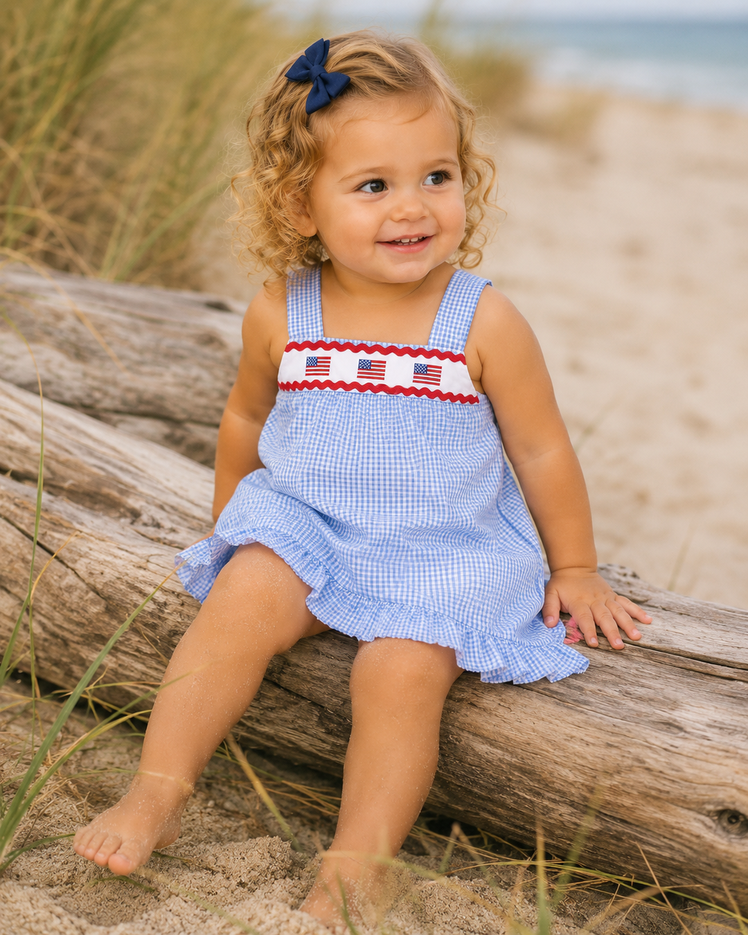 Child in a blue dress with American flag details sitting on driftwood at the beach.