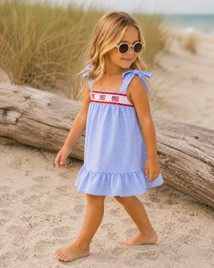 Young girl in a blue dress with an American flag design, standing on a sandy beach.