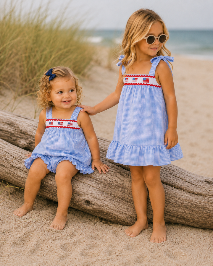 Two young girls in matching blue dresses with American flag details on a beach.