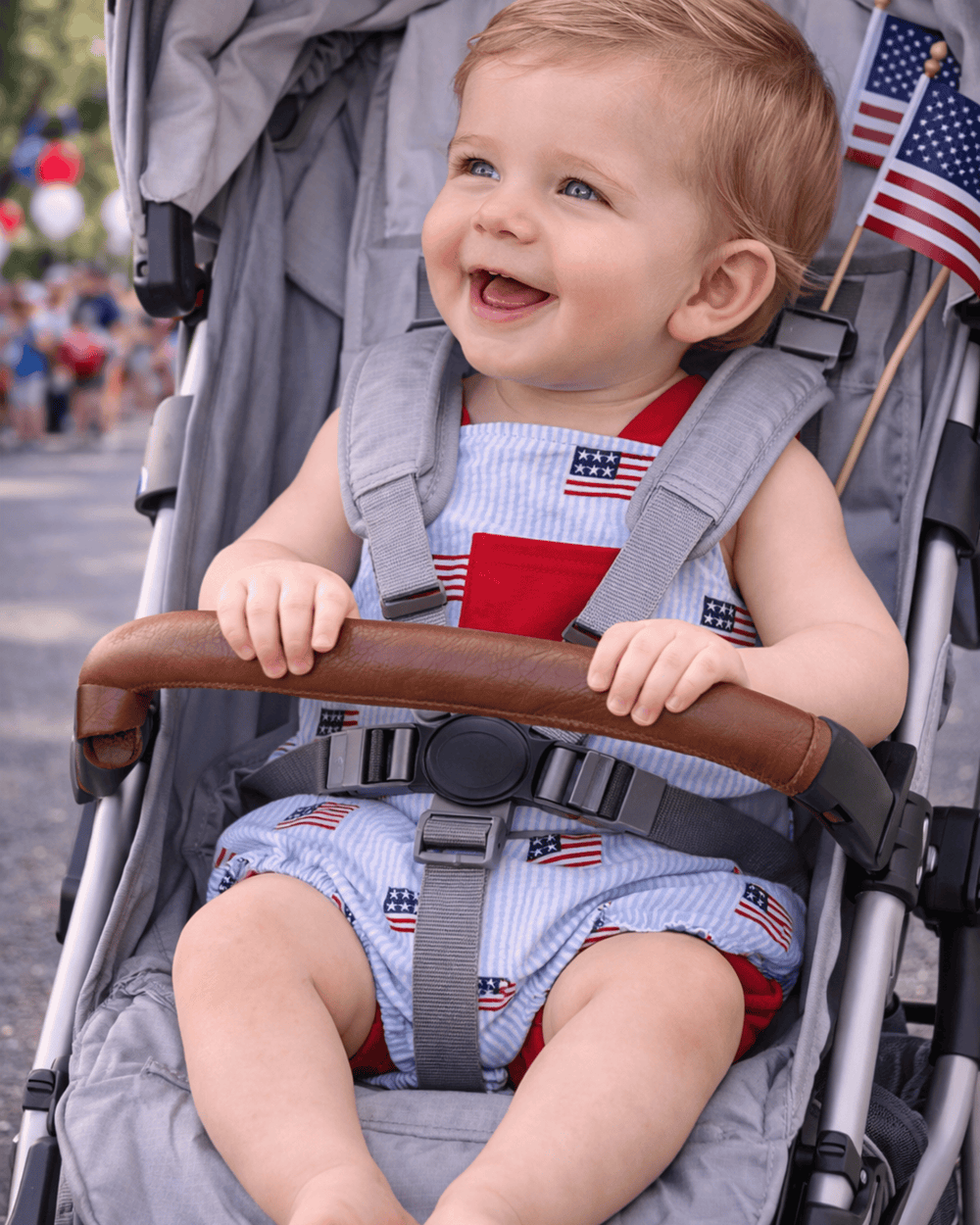 Child in a stroller wearing a outfit with American flag patterns, smiling.