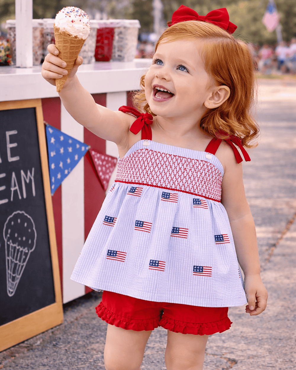 Child in a patriotic outfit holding an ice cream cone in front of an ice cream stand.