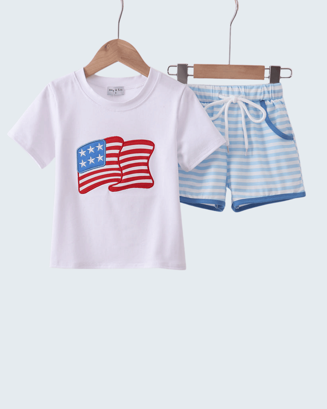 White t-shirt with American flag design and blue striped shorts on hangers against a white background