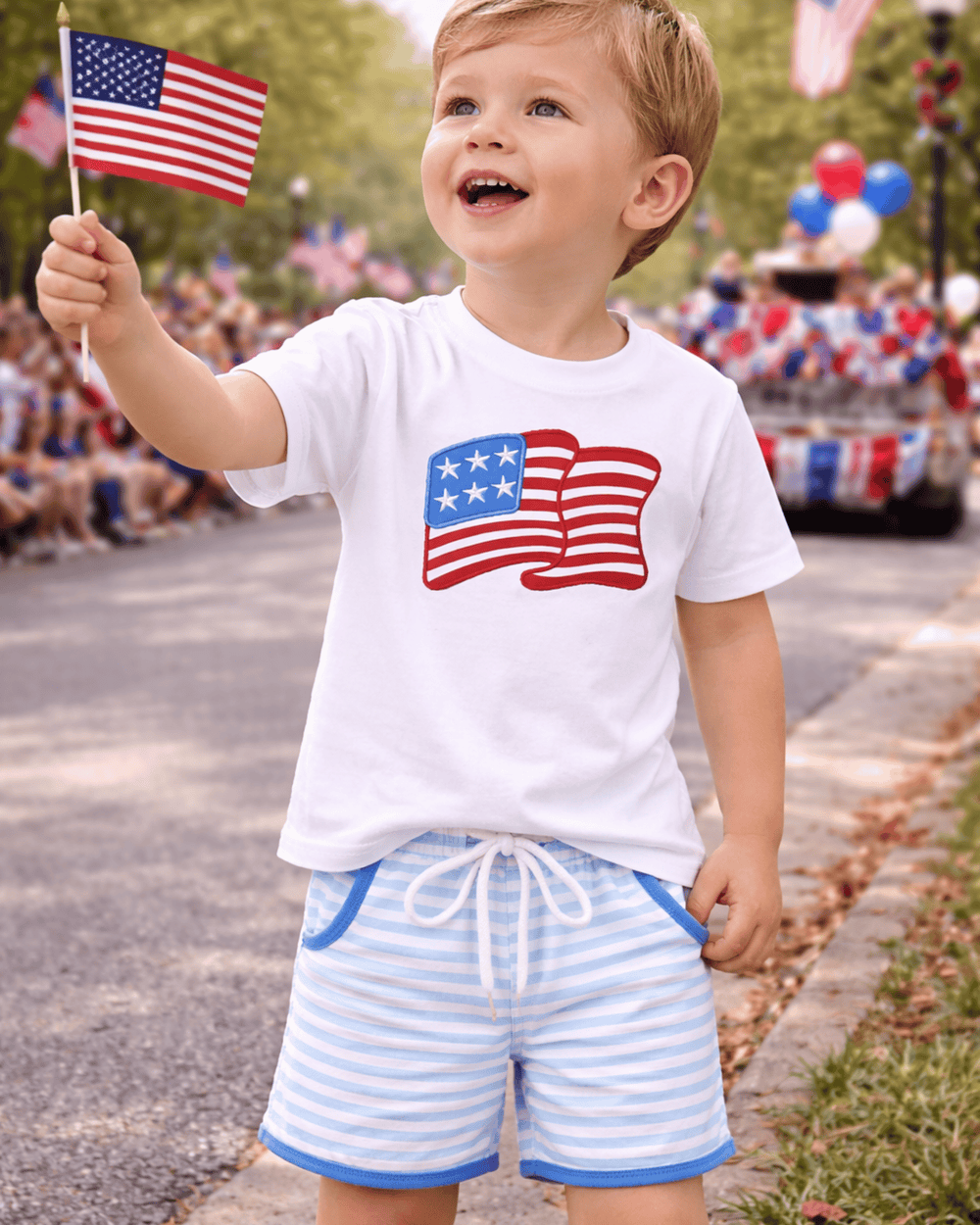 Child wearing a white t-shirt with an American flag design and blue shorts, holding a small American flag, standing in front of a parade.