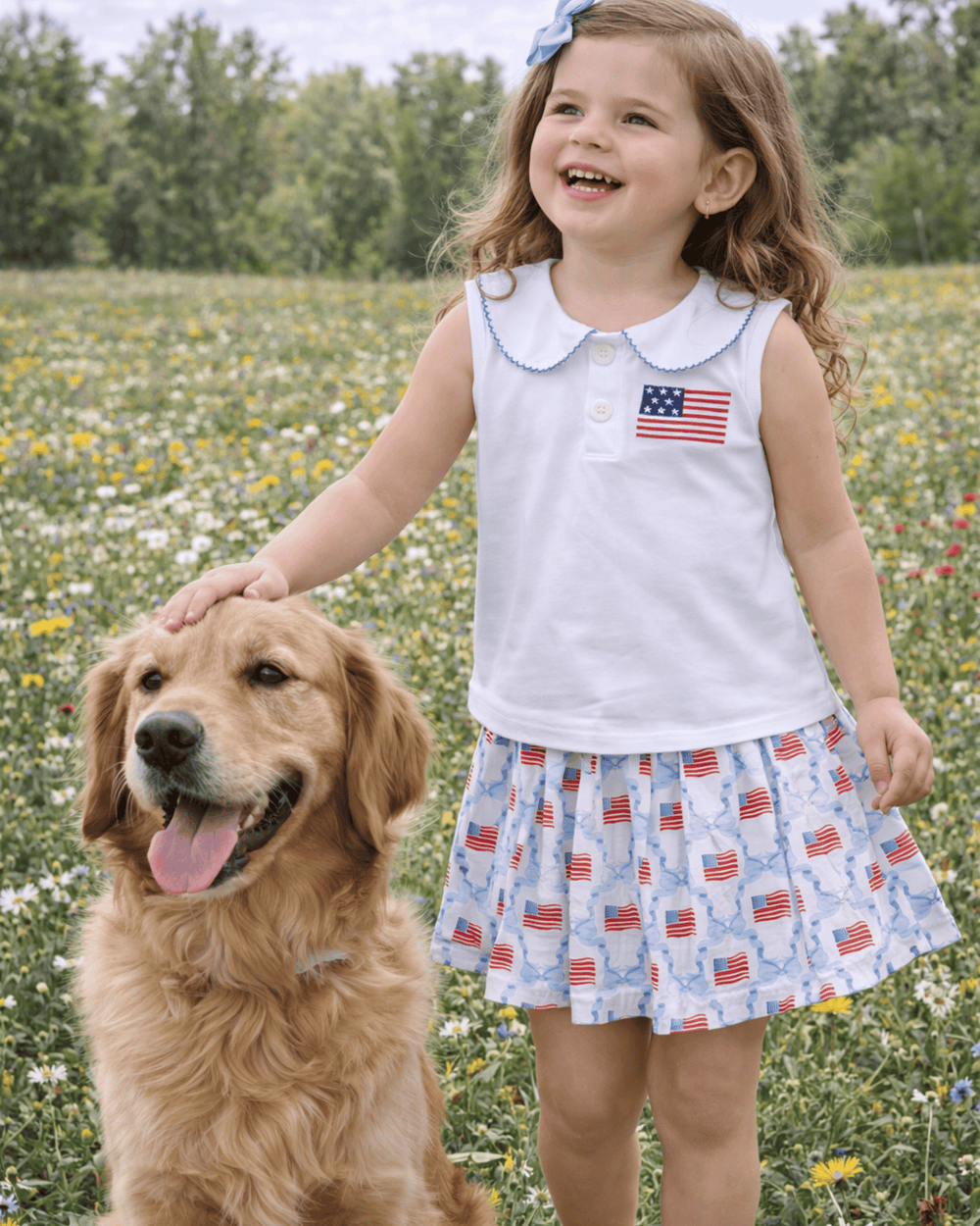 Young girl in a field with a dog, wearing a dress with an American flag design.