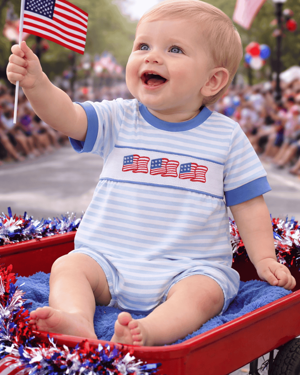 Baby in a red wagon holding an American flag, wearing a onesie with American flag design.