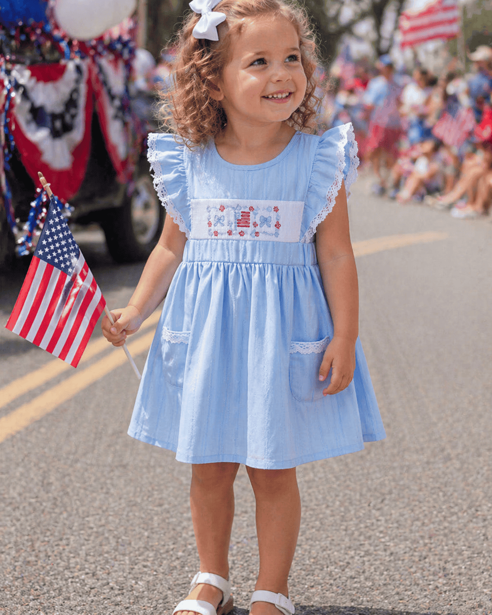 Young girl in a blue dress with ruffles and American flag design, holding a small American flag, in a parade setting.