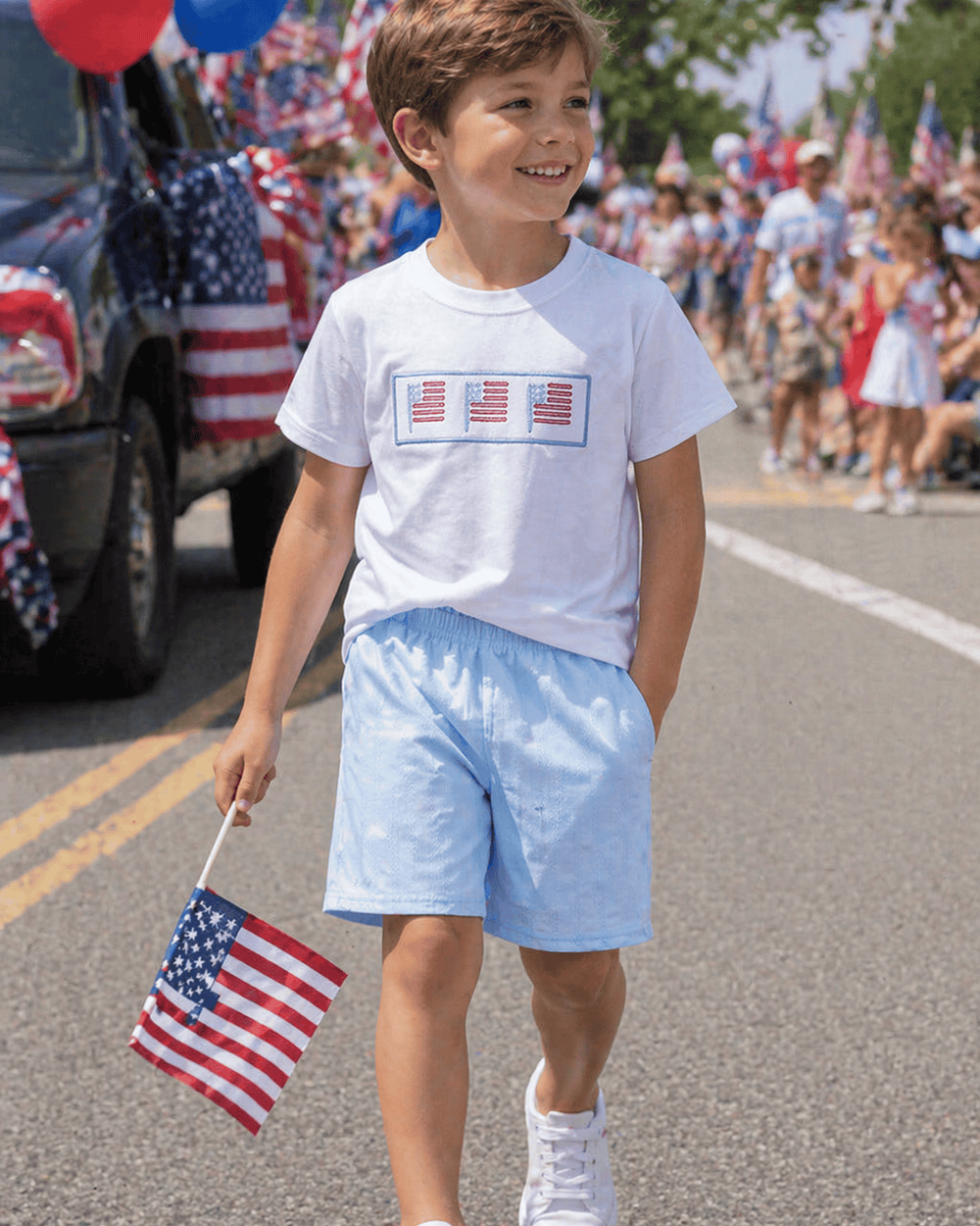 Young boy in a parade holding an American flag, wearing a white t-shirt with flag design and light blue shorts.