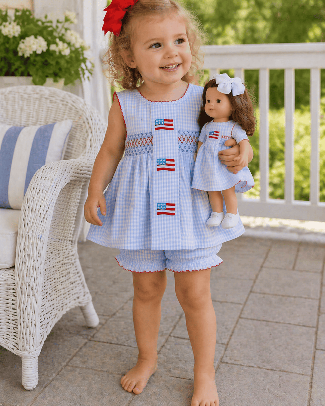 Child wearing a matching dress with American flag patterns, holding a doll in a similar outfit, on a patio.