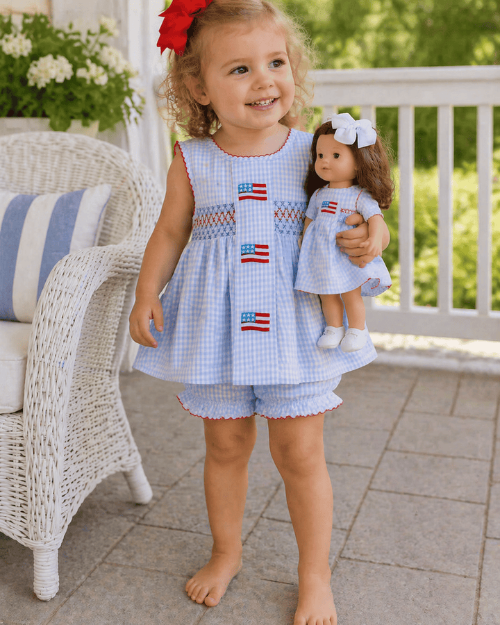 Child wearing a matching dress with American flag patterns, holding a doll in a similar outfit, on a patio.