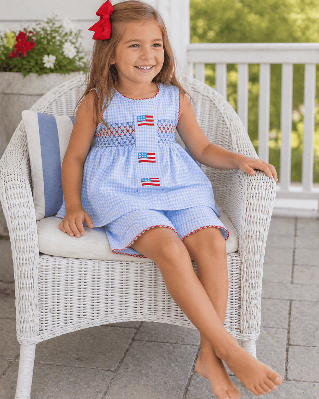 Young girl sitting on a white wicker chair wearing a blue dress with American flag patterns.