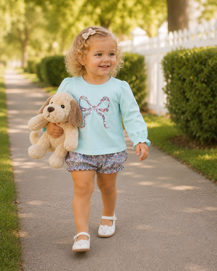 A young toddler girl walking on a sunny outdoor path, holding a plush dog toy. She is wearing a light blue long-sleeve top with a glittery bow graphic on the chest, patterned ruffled shorts, and white Mary Jane shoes.