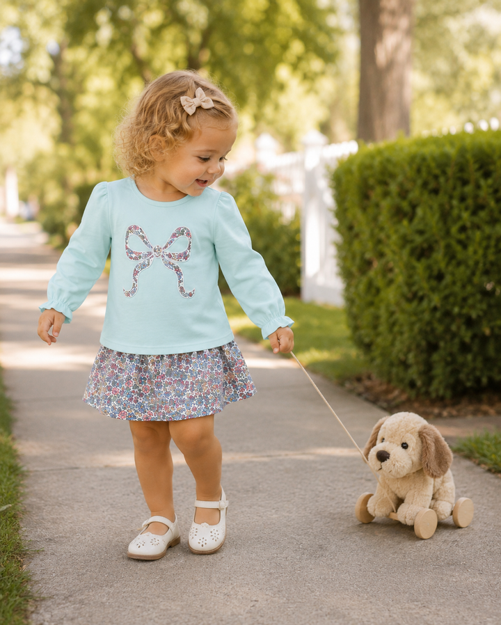 A toddler girl walking on a paved path outdoors, wearing a light blue long-sleeve top with a bow graphic and a floral skirt, while pulling a small plush dog toy.