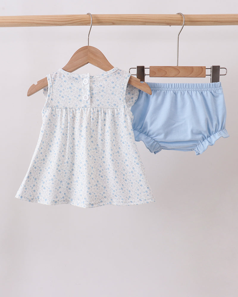 White baby dress with floral pattern and light blue bloomers hanging on a wooden rack against a plain background.