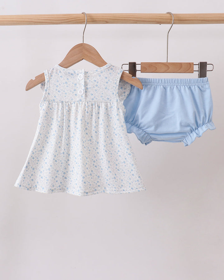 White baby dress with floral pattern and light blue bloomers hanging on a wooden rack against a plain background.