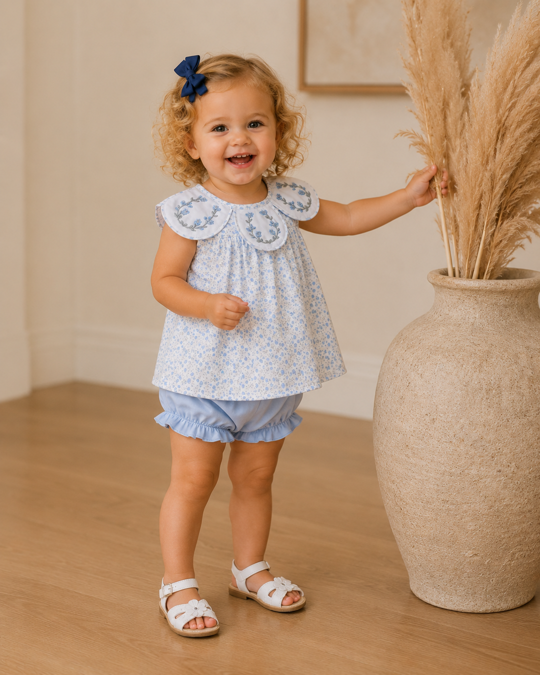 Child in a floral top and shorts standing next to a vase with dried plants indoors.