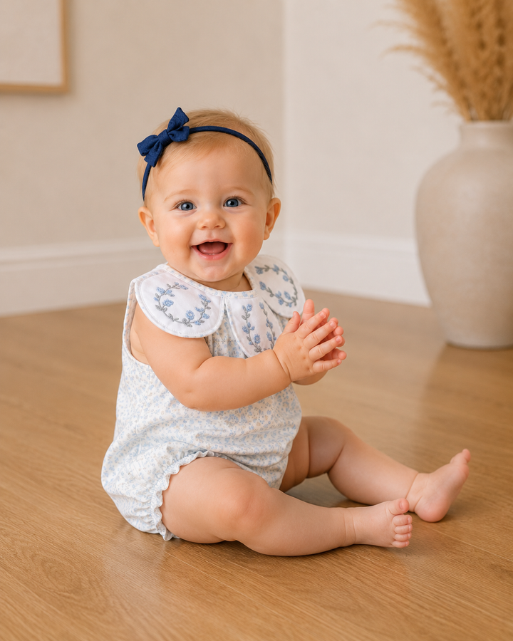 Baby sitting on a wooden floor wearing a white lace romper with a blue bow headband.
