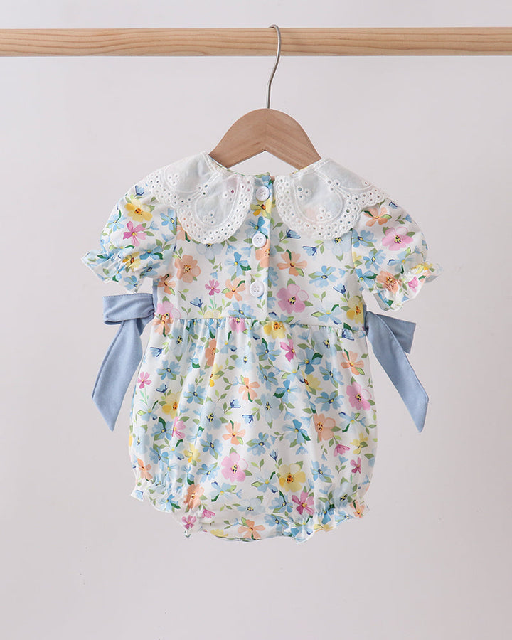 Floral baby romper with lace collar and blue ribbons on a wooden hanger against a light gray background